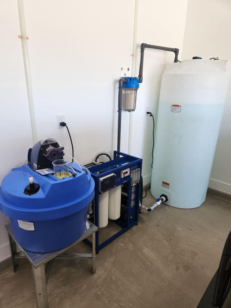 A commercial reverse osmosis skid with membrane housings and a control panel, standing between a blue chemical feed drum and a large white bulk storage tank.