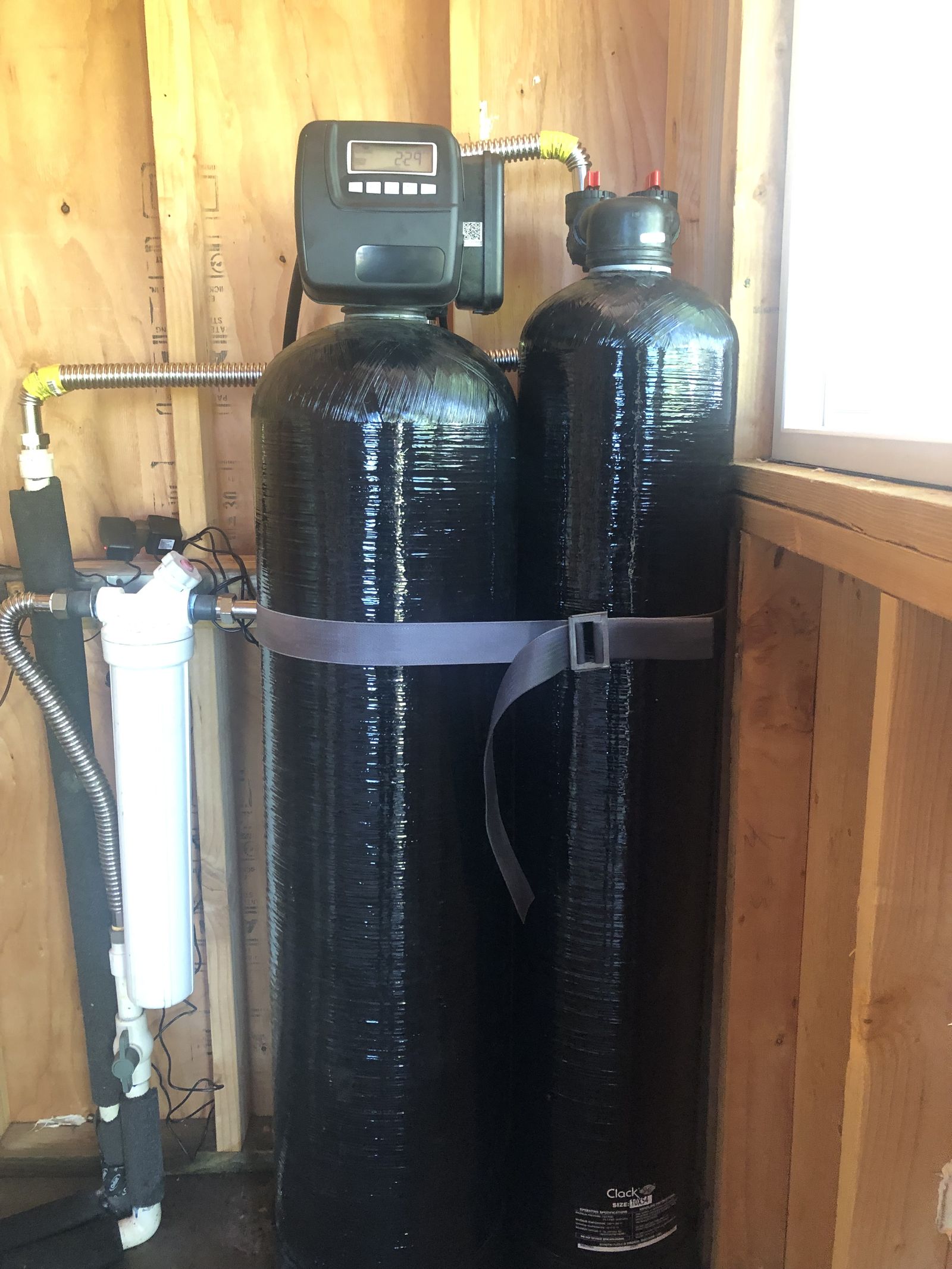 Two black media tanks strapped side by side under a timber floor, joined by braided connectors to a metered control valve.