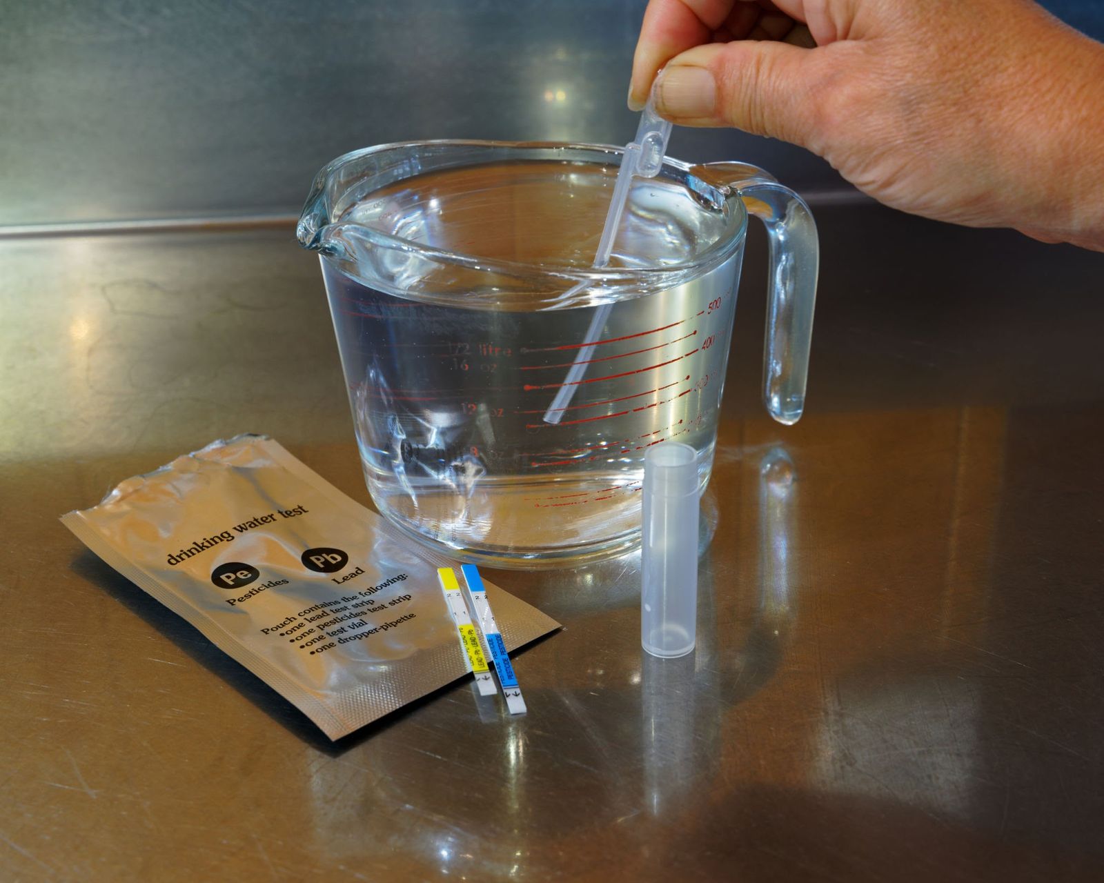 A hand dipping a test strip into a glass jug of water on a steel counter, beside a drinking water test kit for lead and pesticides.