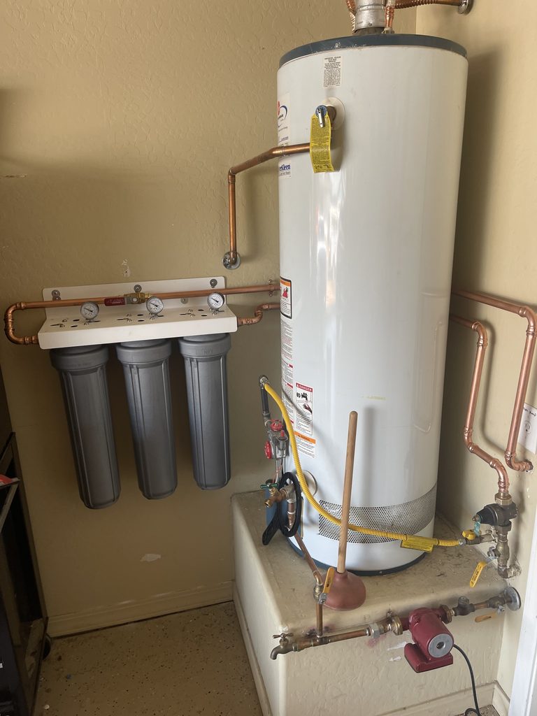 A close view of copper piping crossing the top of a gas water heater to a red circulation pump and shutoff valves on the adjacent wall, beside a labeled electrical outlet.